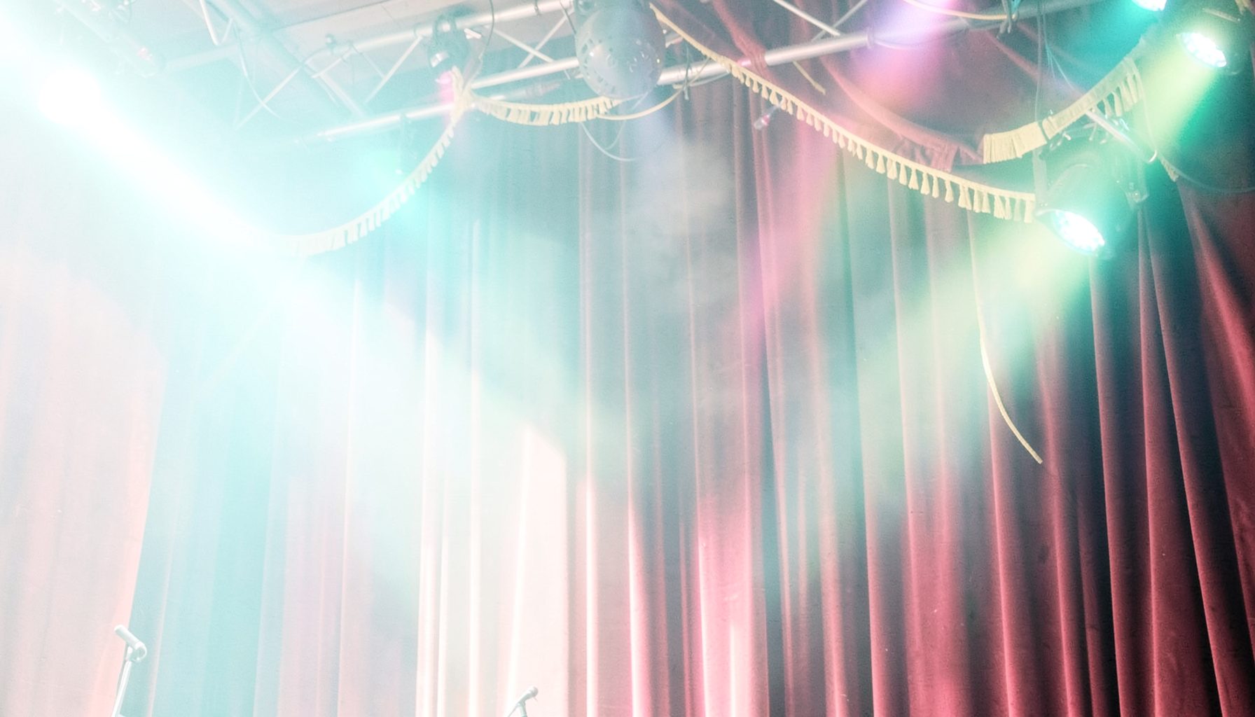 A bright, airy scene of colorful stage lights and a velvet curtain in deep red and gold tones, evoking the excitement of live entertainment