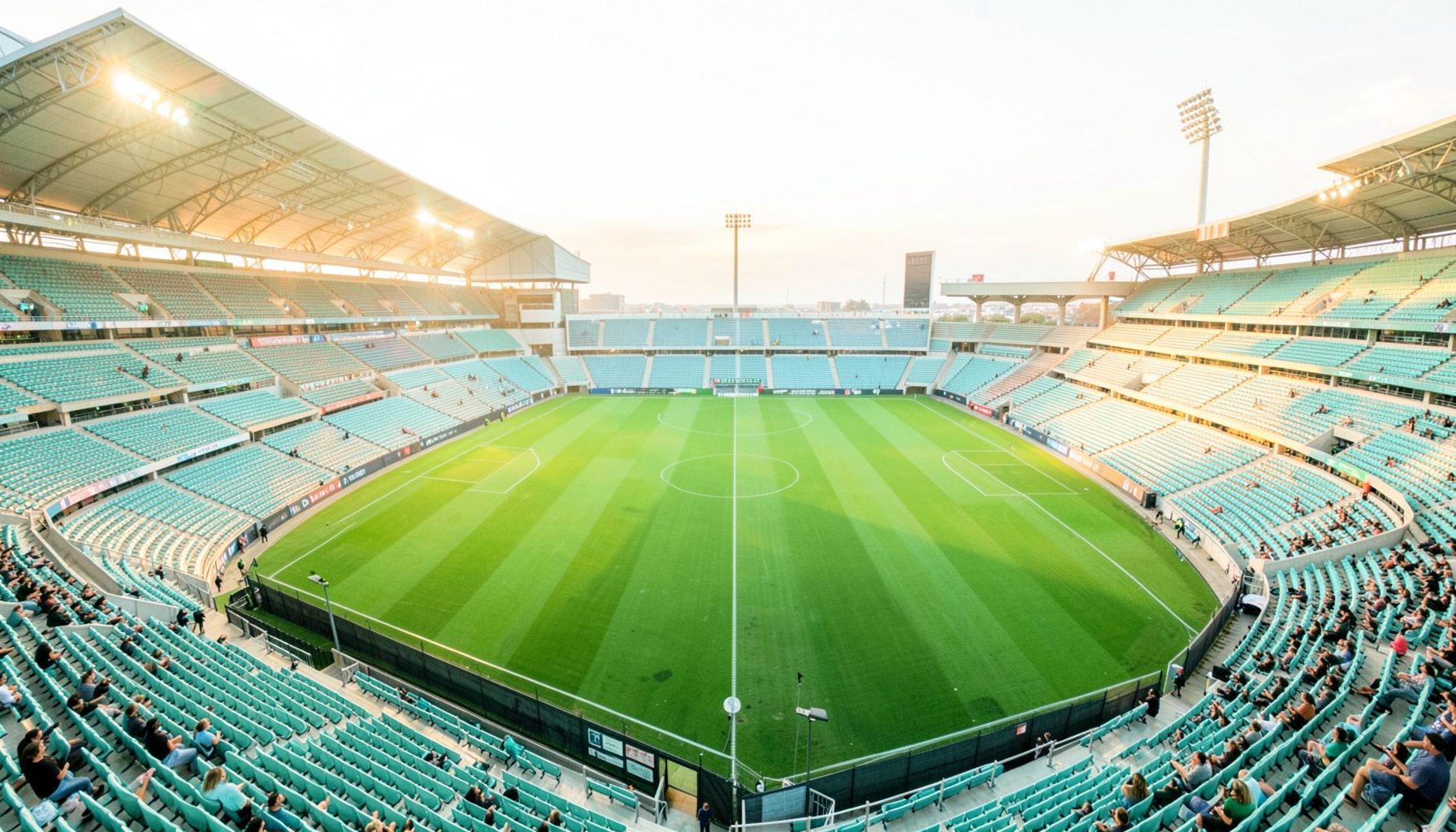 Wide aerial view of a brightly lit sports stadium at dusk, green field and empty seats bathed in warm golden light, calm anticipation