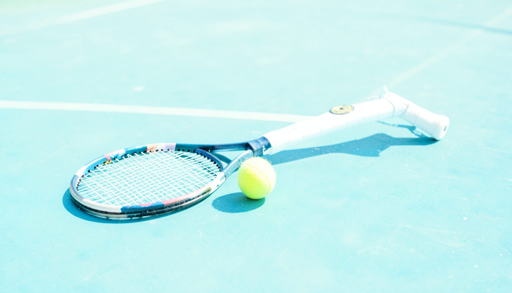 Tennis racket and yellow ball on a blue hard court, bright midday sun casting sharp shadows, competitive energy