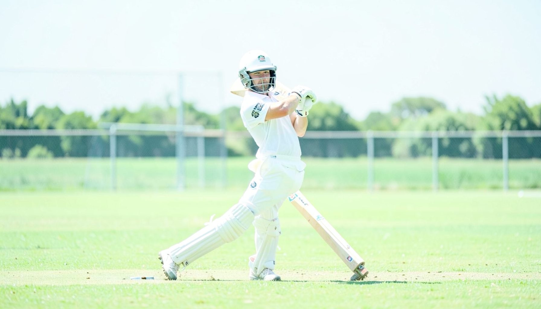 Cricket batsman in white uniform playing a cover drive, green field stretching to the boundary, bright summer afternoon