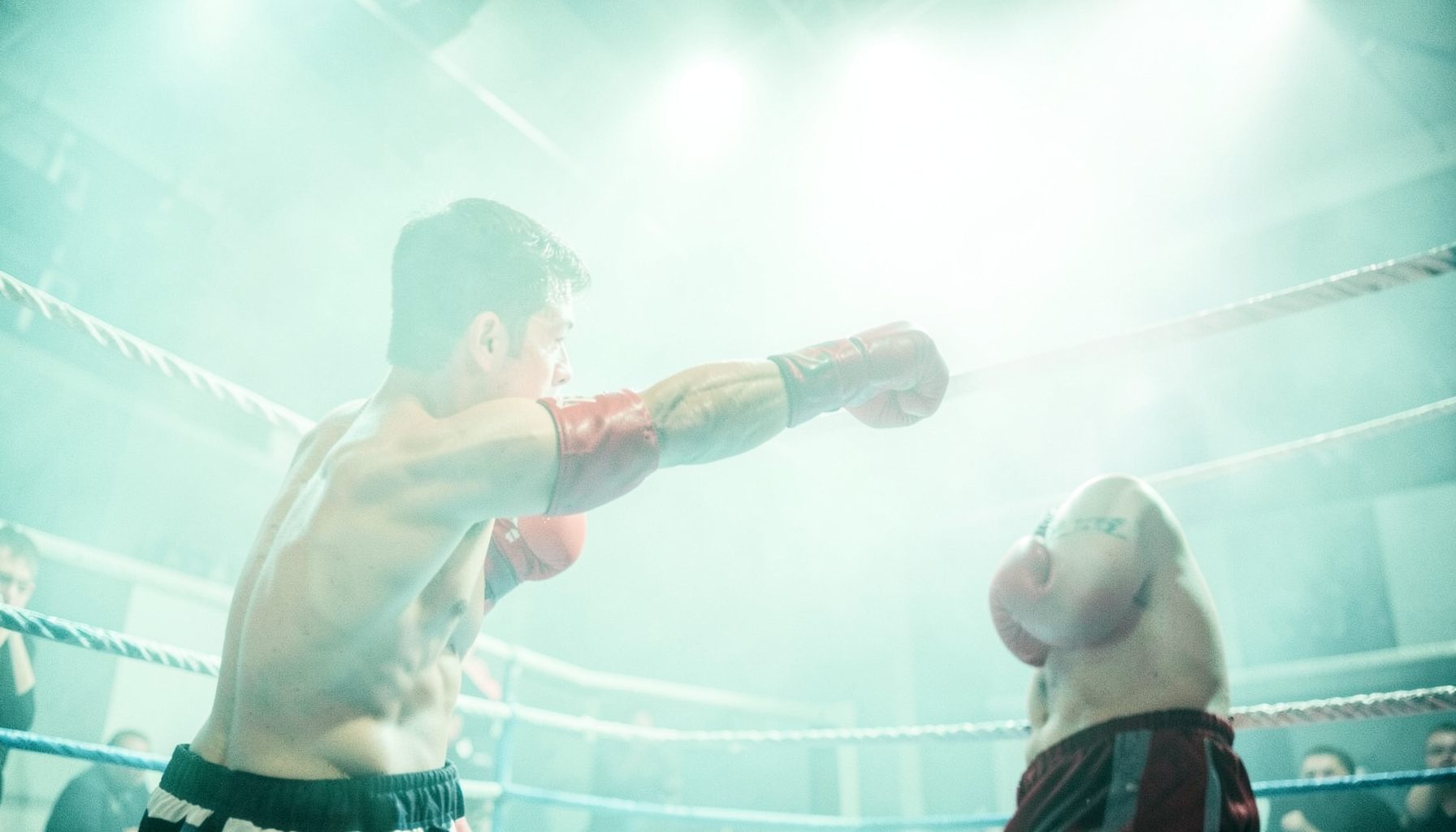 Boxer in red gloves throwing a jab inside a ring, ropes visible, dramatic overhead lighting cutting through haze
