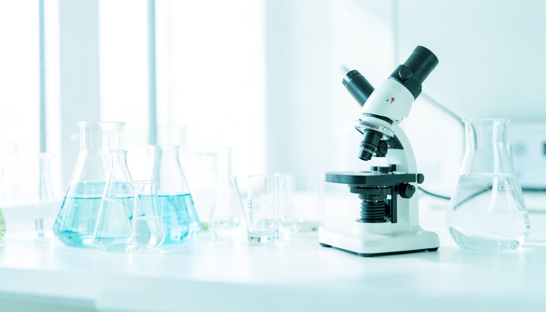 A bright high-key photograph of a modern laboratory bench with glass beakers and a microscope, bathed in soft natural light with clean white and pale blue tones.