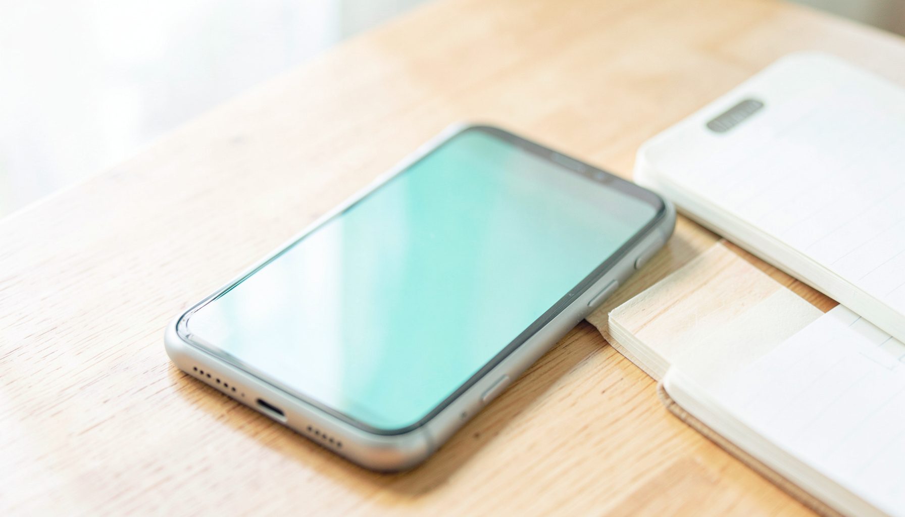 A smartphone resting on a wooden desk beside a notebook, soft natural daylight streaming in, warm wood and white tones
