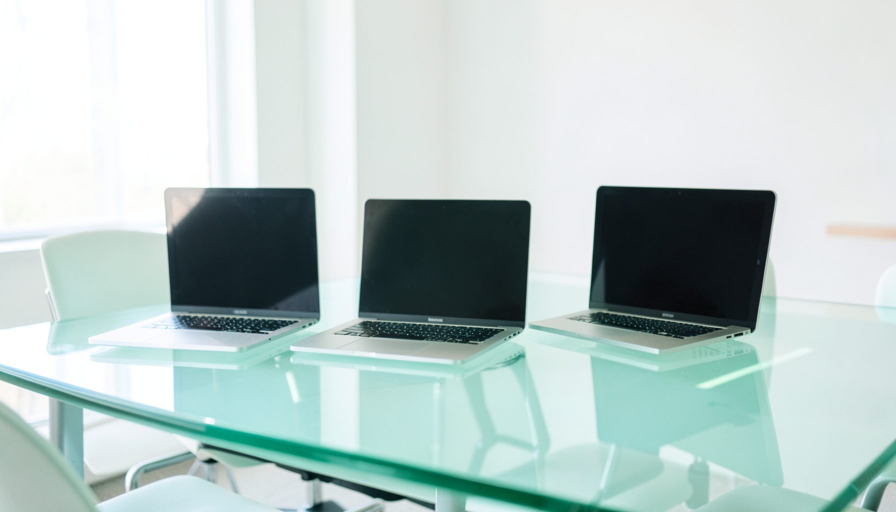 Three sleek laptops on a glass conference table, bright office setting with natural light and white walls